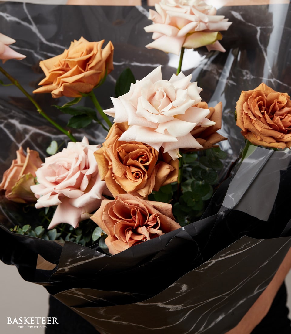 A lavish bouquet of roses elegantly wrapped in chic black and white marbled paper. The exquisite blooms feature a harmonious blend of peach, pale pink, and rust hues. In the corner, a sophisticated Basketeer logo with refined text underneath graces the scene, adding an extra touch of luxury. The backdrop is minimalist, allowing the bouquet's opulence to truly shine.