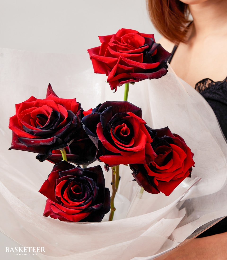 A woman in an elegant black top gracefully holds a luxurious bouquet of five stunning red roses, each uniquely edged with black for a dramatic effect. The exquisite arrangement is wrapped in sheer white paper, adding to its sophisticated allure. The prestigious Basketeer logo subtly enhances the scene from the bottom left corner, signifying the essence of opulent gifting.