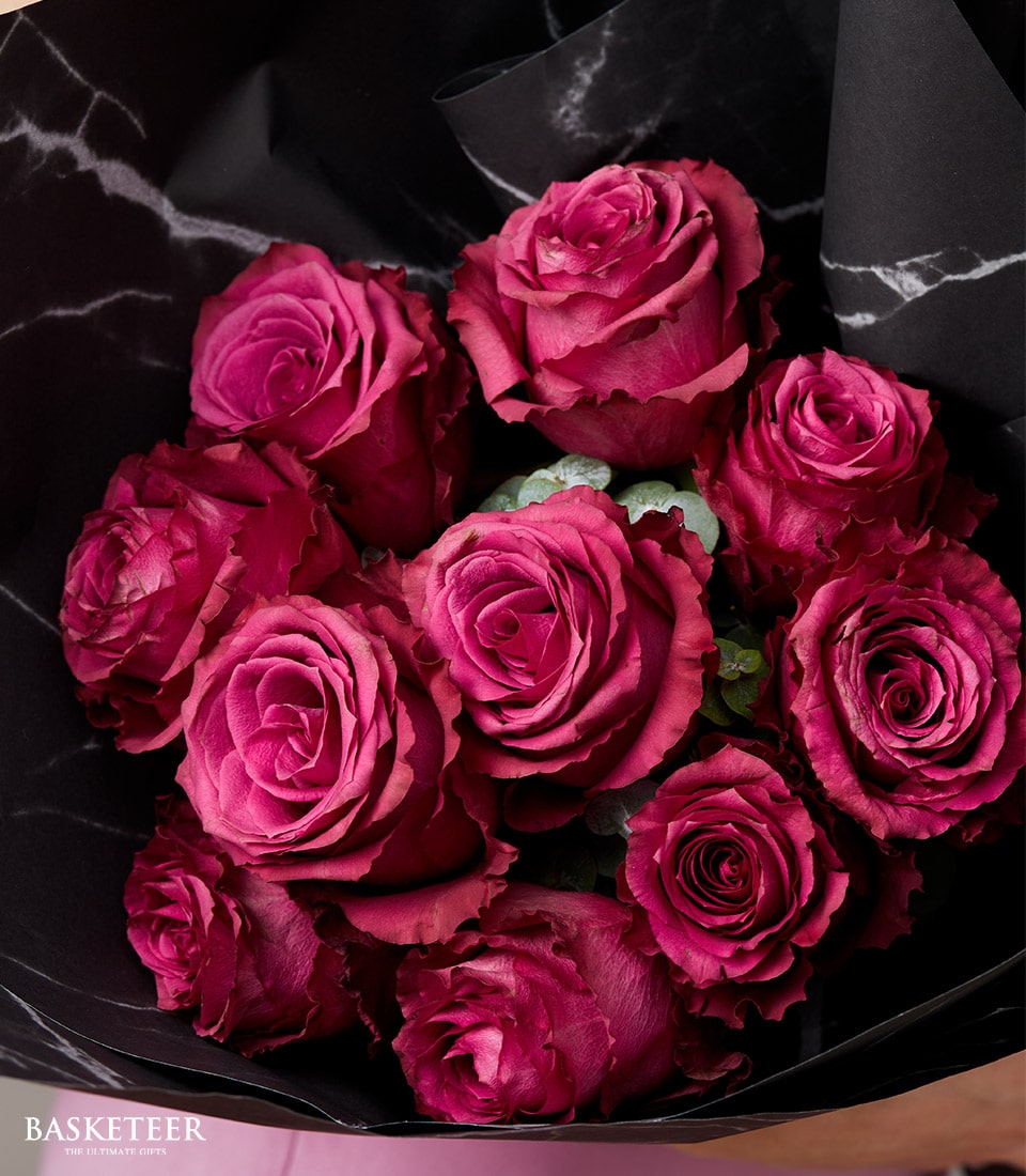 An exquisite bouquet of opulent dark pink roses, meticulously arranged against a sophisticated black backdrop. The lush, velvety petals of the vibrant roses are intricately clustered together, forming a captivating visual masterpiece that is further enhanced by the elegant wrapping. At the base, the prestigious 