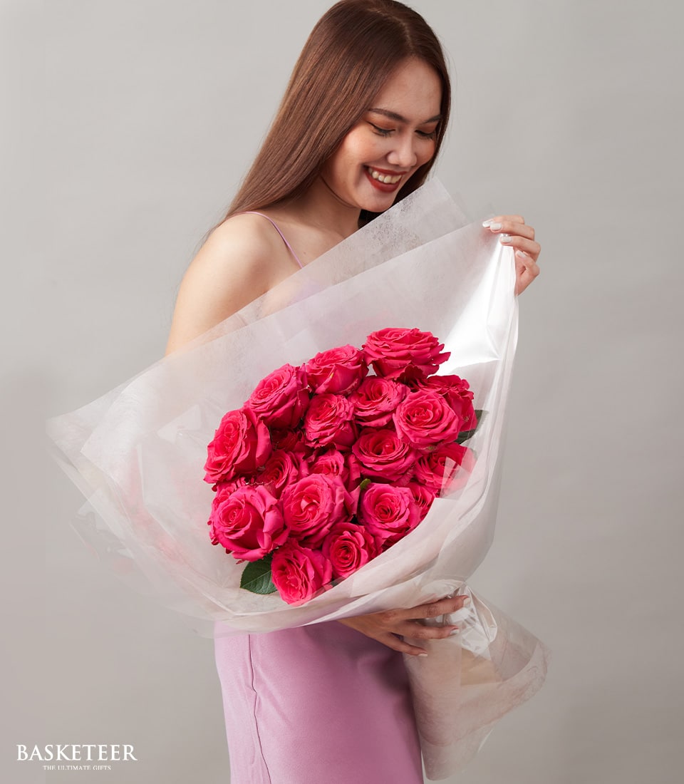 A radiant woman with flowing brown hair beams with joy as she cradles the luxurious Ultimate Rose Spectacle Bouquet, elegantly wrapped in pristine white paper. Clad in a delicate light pink sleeveless dress, she stands poised against a sophisticated neutral backdrop. The image is graced with the prestigious 