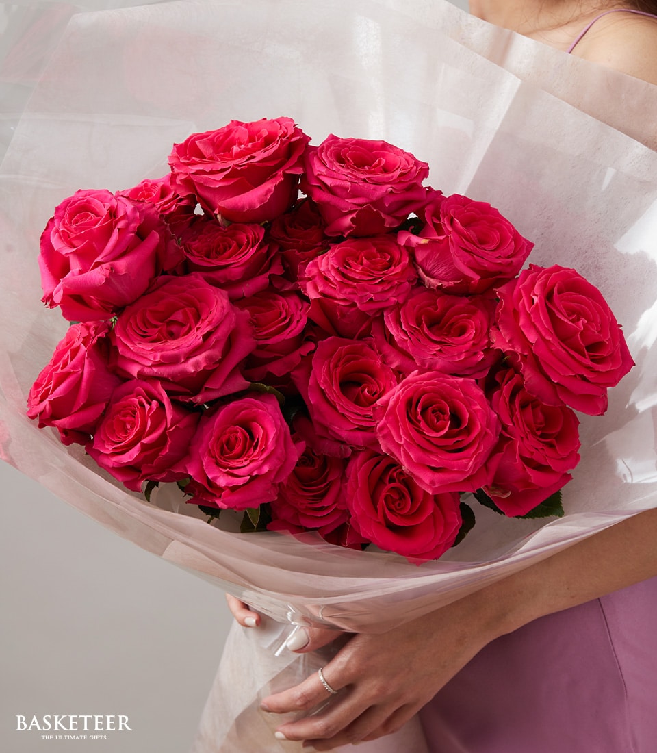 A person in an elegant pink dress gracefully holds The Basketeer’s Ultimate Rose Spectacle Bouquet, a stunning arrangement of lush red roses wrapped in pristine white paper. Each rose is meticulously placed to create a harmonious and vibrant display. Peekings of the luxurious Basketeer logo ribbon add an exquisite touch, enhancing the allure of this captivating floral masterpiece. Only the lower part of the person's face and body are partially visible, drawing full attention to the opulent bouquet they cradle with care.