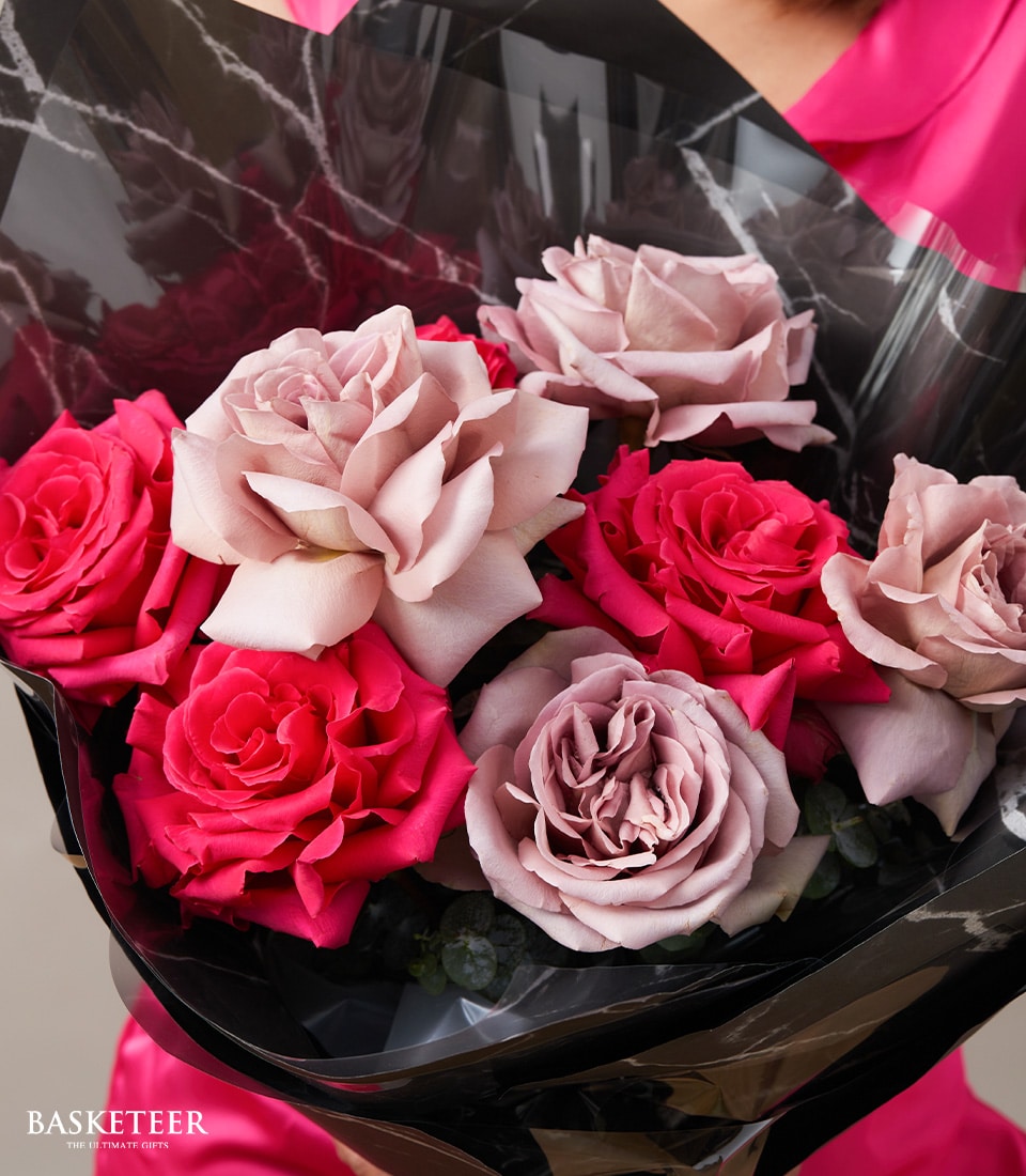 A luxurious bouquet of vibrant pink and delicate pale mauve roses, elegantly wrapped in sophisticated black floral paper. The stunning arrangement is cradled by a person adorned in a chic, bright pink top, perfectly complementing the blossoms. In the bottom left corner, the prestigious Basketeer logo reads 