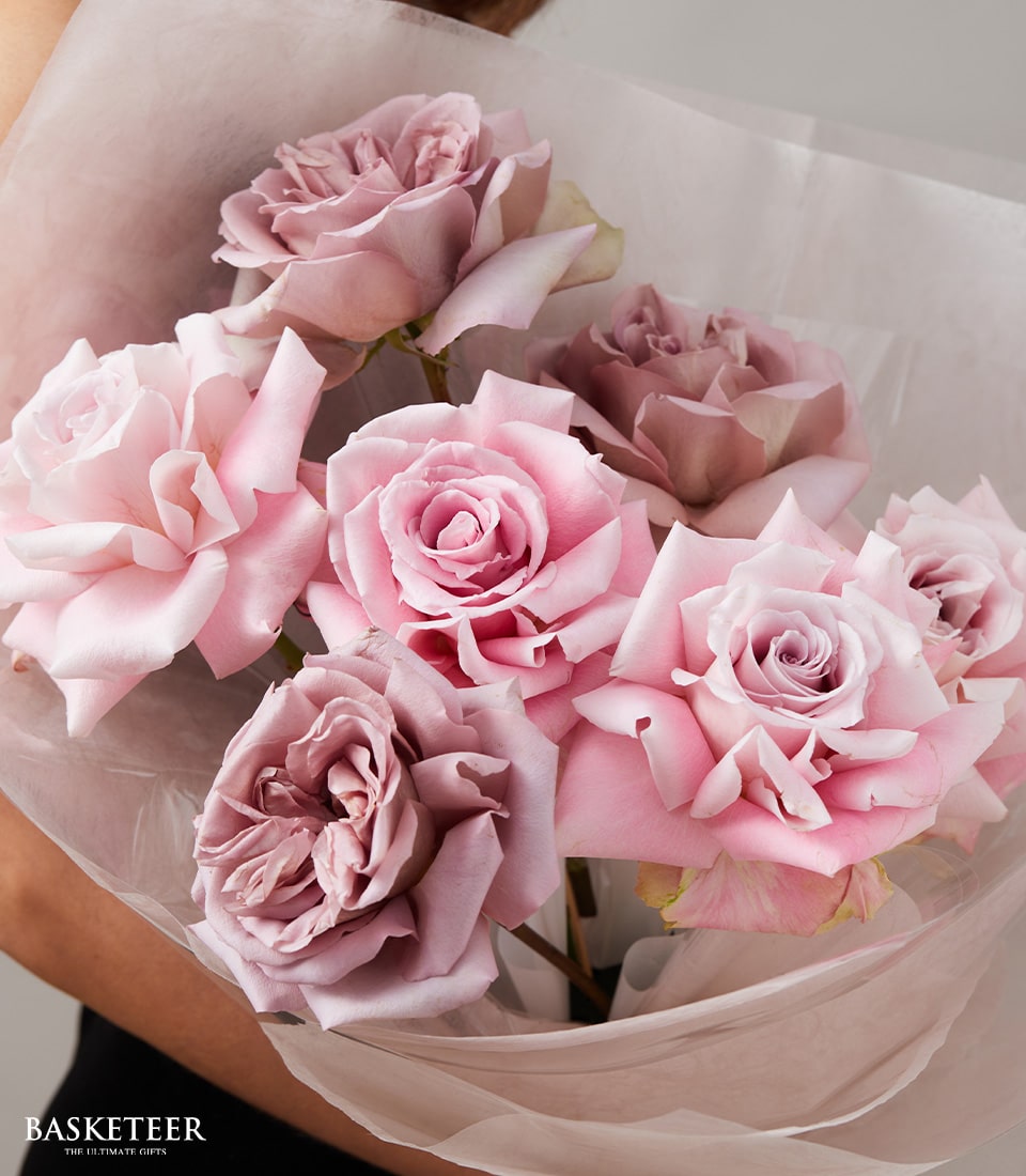 A pair of elegant hands tenderly cradles a sumptuous bouquet brimming with exquisite roses in delicate hues of pink and lavender, impeccably wrapped in silky translucent white paper. Positioned gracefully at the bottom left, the sophisticated logo and text 