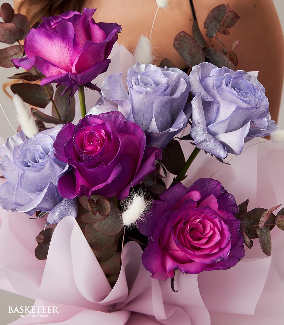 A sumptuous bouquet featuring an array of luxurious purple, pink, and lavender roses interspersed with delicate eucalyptus leaves and soft white accents. The exquisite flowers are meticulously wrapped in a chic light pink paper, enhancing their fresh and vibrant allure. The brand 