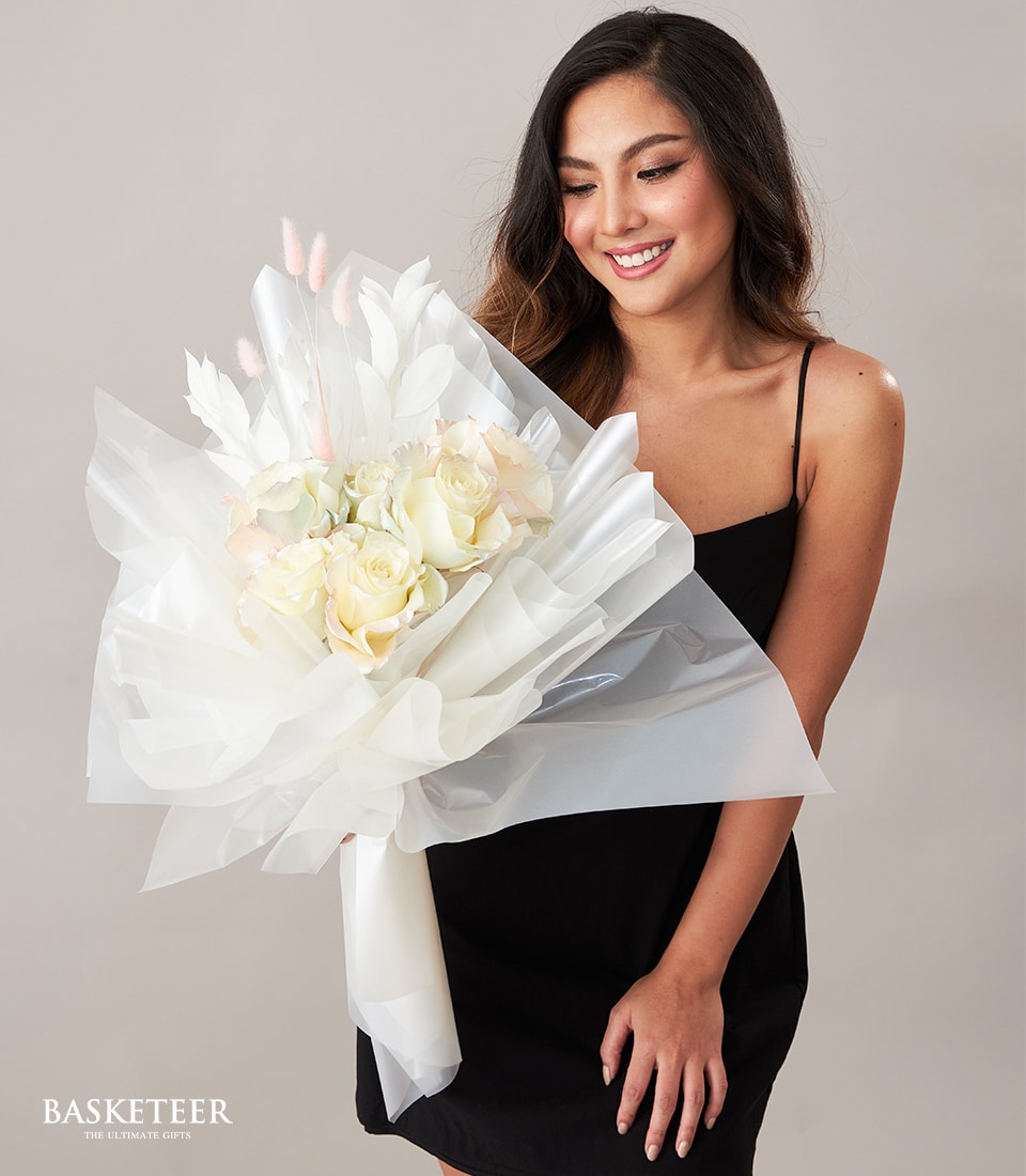 A sophisticated woman with long, flowing dark hair, elegantly clad in a black sleeveless dress, beams with joy while cradling a sumptuous bouquet of white and pale pink flowers wrapped in delicate translucent white paper. The prestigious Basketeer logo proudly graces the pristine wrapping, accompanied by the eloquent phrase 