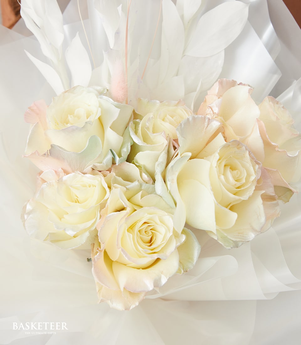 A close-up of a lavish bouquet featuring exquisite pale cream and soft pink roses, meticulously nestled within translucent white wrapping paper. The arrangement exudes elegance and delicacy, with the soft texture and subtle color variations of the petals enhancing its luxurious appeal. A ribbon adorned with the Basketeer logo adds a refined touch to this sophisticated floral masterpiece.
