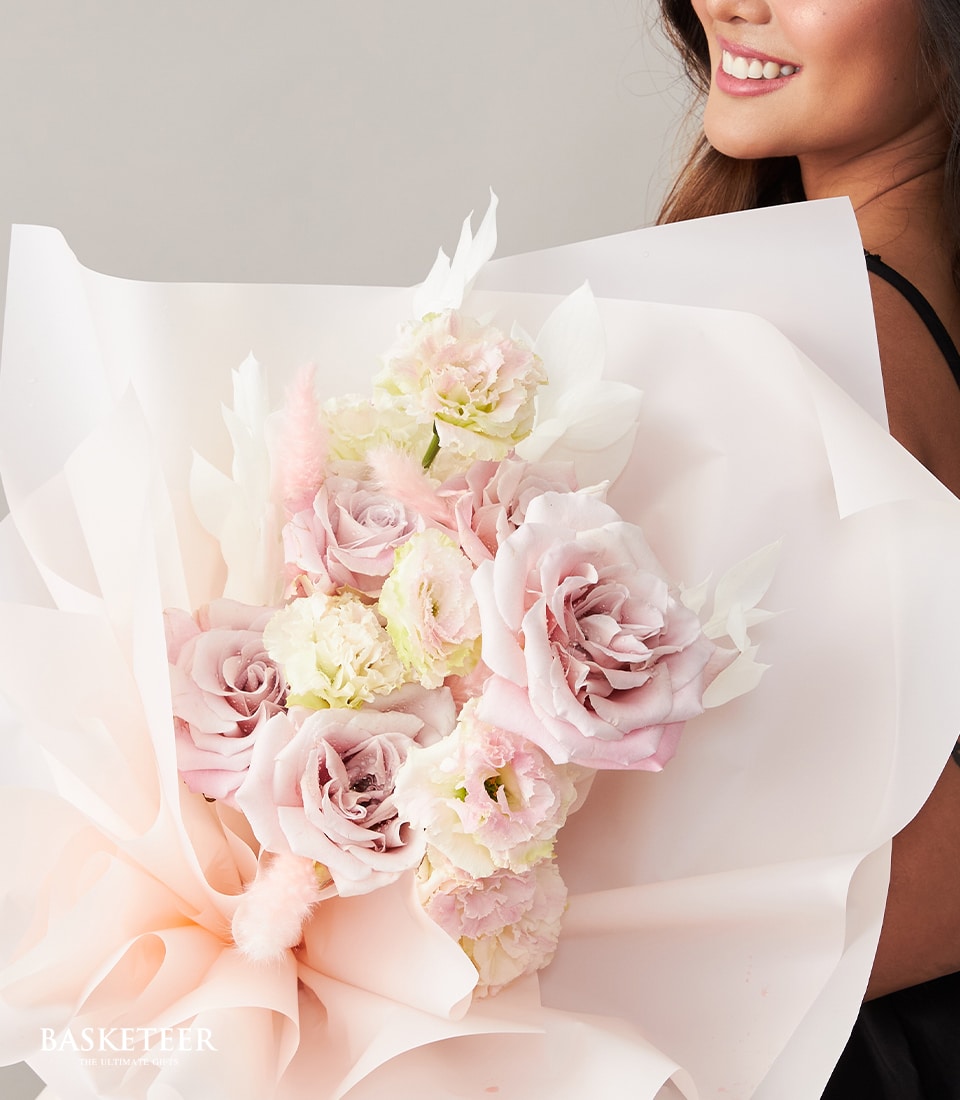 A person with long, dark hair beams with joy while cradling an opulent bouquet of pastel-colored roses and an exquisite assortment of blooms, elegantly wrapped in delicate pink and white paper. Adorned with the Basketeer logo on a ribbon, the luxurious arrangement exudes sophistication against a plain, light-toned background.