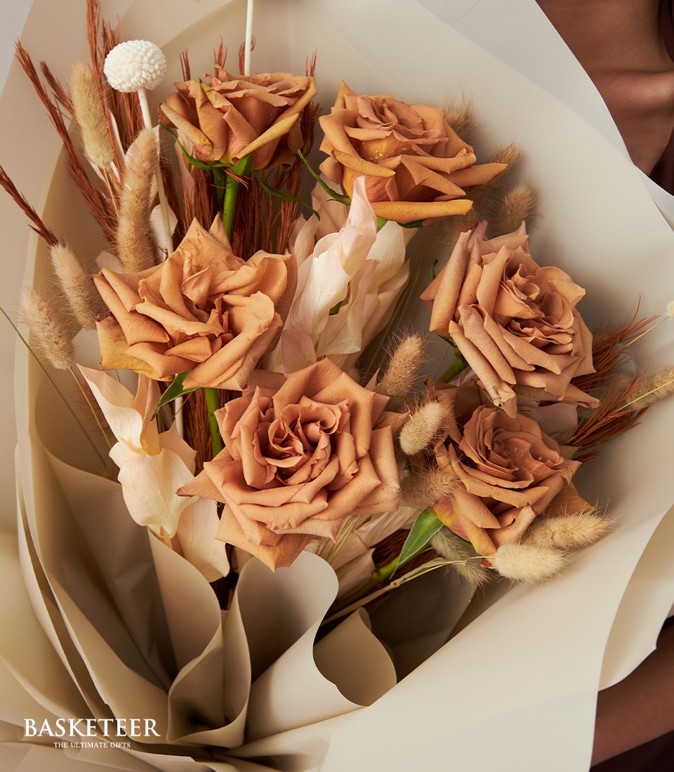 A lavish bouquet of elegantly dried, golden-brown roses complemented by a delicate array of beige and ivory filler flowers. The arrangement is tastefully wrapped in sophisticated white and light beige paper, exuding a modern minimalist charm. Proudly adorned with the prestigious Basketeer logo in the bottom left corner, this scene invites an essence of refined luxury and captivating warmth.