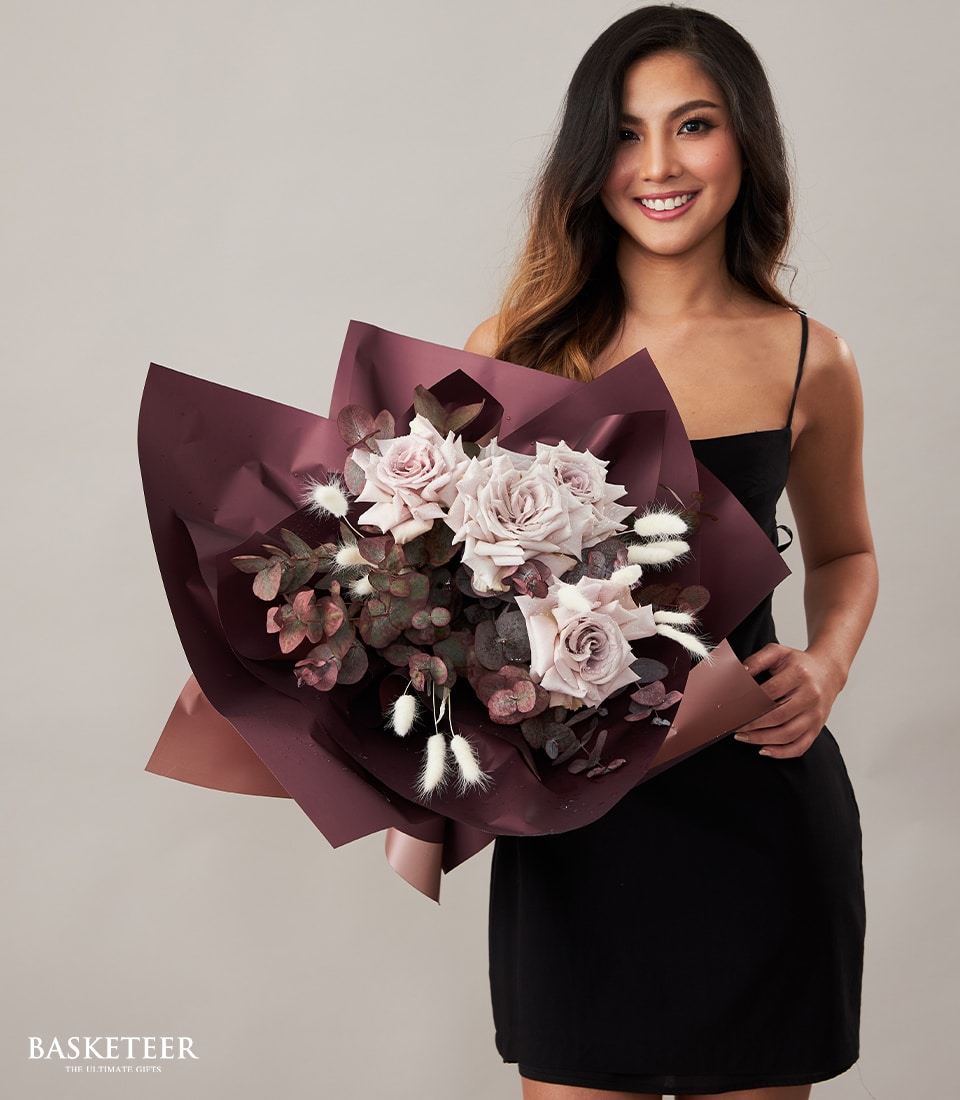 A radiant woman with long, flowing dark hair elegantly holds a luxurious bouquet featuring soft pink roses, rich dark green foliage, and delicate white dried flowers. She is draped in a sophisticated black dress. A chic ribbon adorned with the Basketeer logo gracefully encircles the bouquet. In the bottom left corner, the distinguished text “BASKETEER – The Ultimate Gift” makes its presence known, adding an air of exclusivity to this enchanting scene.