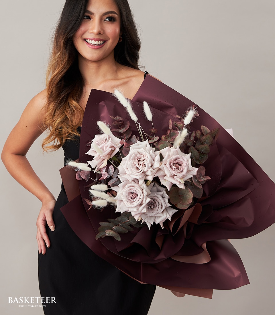 A person with flowing long hair, elegantly attired in a chic black ensemble, is beaming with delight while cradling an exquisite bouquet of pale lavender roses wrapped in sumptuous deep burgundy paper. The arrangement is tastefully accented with delicate dried foliage and soft white bunny tail grass, exuding luxury and sophistication. In the bottom left corner, the prestigious 