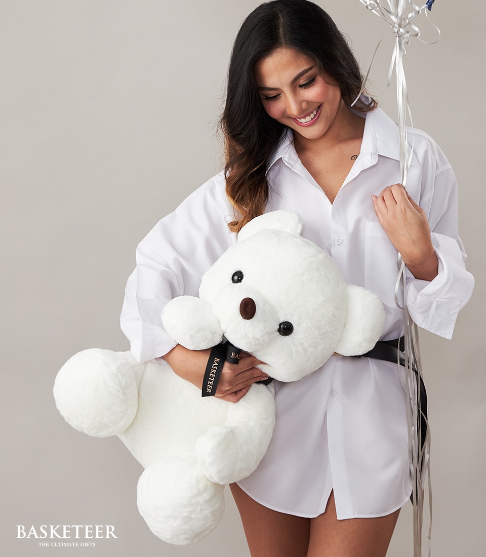 A woman in an elegant oversized white shirt exudes joy as she embraces a sumptuous, large white teddy bear and a dazzling bouquet of silver and blue balloons. The scene is set against a sleek gray background, where the sophisticated Basketeer logo with the tagline 