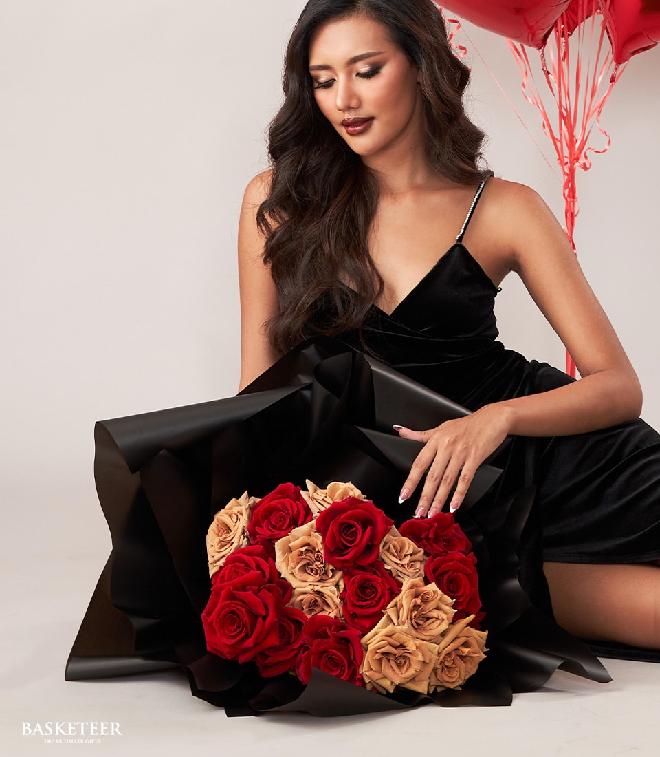 A woman in an elegant black dress cradles the opulent Crimson Love Gift Set by Basketeer, showcasing a lavish bouquet of red and peach roses beautifully wrapped in sophisticated black paper. Her hair cascades in loose waves as she gazes tenderly at the exquisite flowers. In the background, luxurious red heart-shaped balloons add to the atmosphere of romance and refinement. A hint of a ribbon with the Basketeer logo subtly enhances this scene, underscoring its elegance and exclusivity.