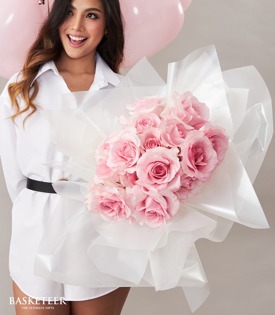 A woman elegantly dressed in a pristine white shirt with a chic black belt cradles an opulent Soft Pink Bouquet, meticulously wrapped in exquisite white paper. She stands poised against a minimalist backdrop adorned with romantic heart-shaped balloons. At the lower left corner, the prestigious 