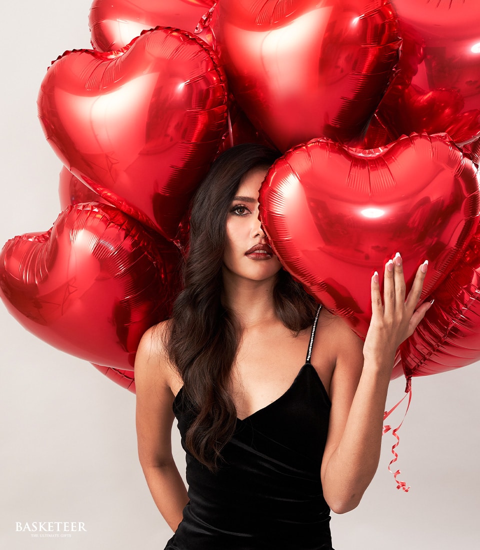 A sophisticated woman with flowing dark hair elegantly graces the scene, holding an exquisite set of vibrant red heart balloons. Dressed in a stunning, sleeveless black dress, she exudes timeless elegance as her hand gently conceals part of her face behind one of the luxurious balloons. The minimalist backdrop ensures that all attention is drawn to her and the lavish Basketeer ribbons adorning the balloons, highlighting their premium quality.