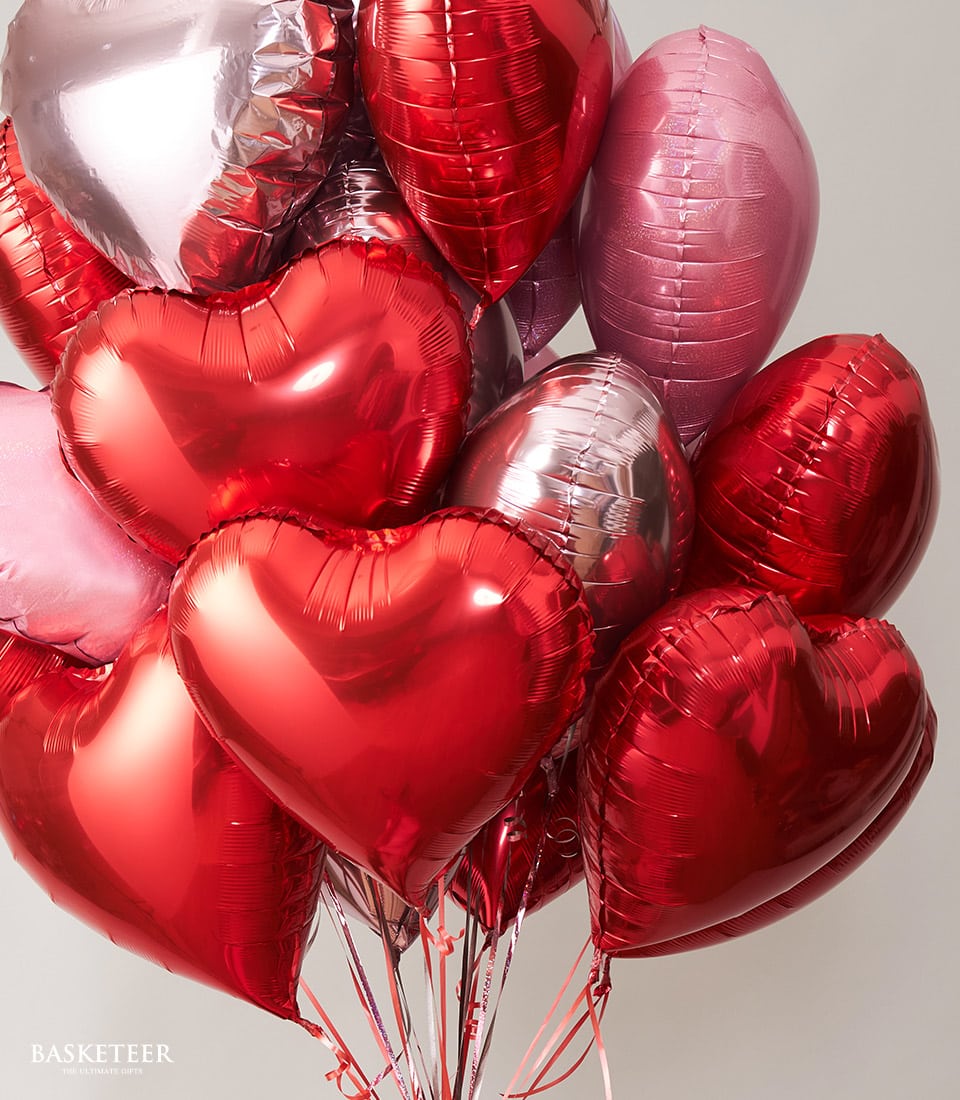 A stunning bouquet of luxurious, heart-shaped balloons in vivid shades of red, pink, and silver elegantly grouped together. Against a sophisticated light gray wall backdrop, these glossy and metallic balloons reflect the light beautifully, creating a radiant and inviting ambiance. Delicate ribbons adorned with the Basketeer logo add an exquisite touch to this opulent scene.