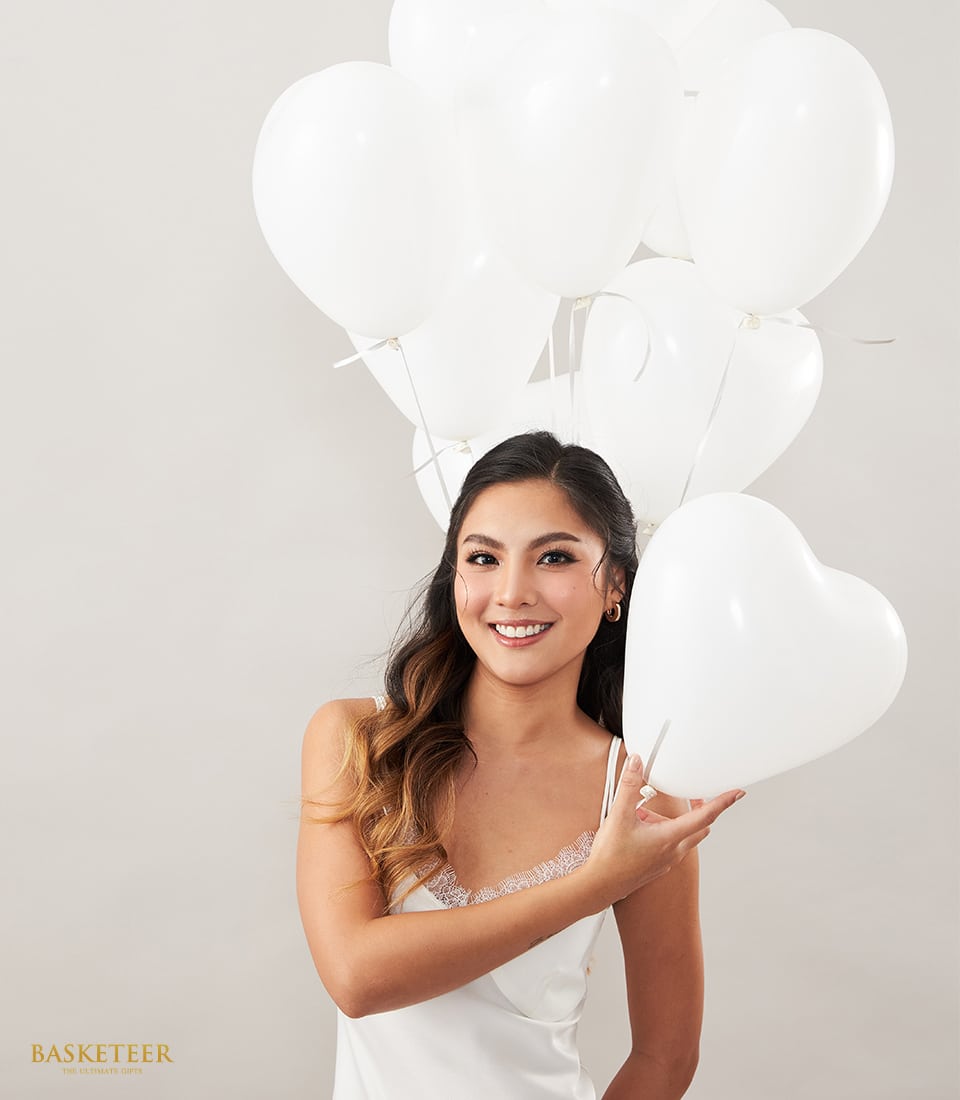 A graceful woman dressed in an elegant white gown beams with joy as she embraces a luxurious bundle of pristine white balloons, including an exquisite heart-shaped one. The backdrop is minimalist and light-hued, exuding sophistication. In the bottom left corner, the esteemed BASKETEER logo adds a touch of exclusivity and refinement to the scene.