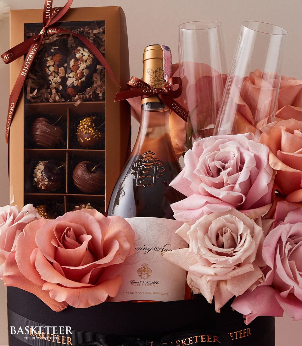 A sumptuous gift arrangement featuring a bottle of fine rosé champagne, paired with two exquisite champagne flutes, awaits your indulgence. A box of decadent assorted chocolates and a breathtaking bouquet of pink and peach roses complete this opulent display. Each item is elegantly adorned with decorative ribbons and meticulously arranged in a luxurious basket from Basketeer, showcasing their signature logo as a mark of unmatched sophistication.