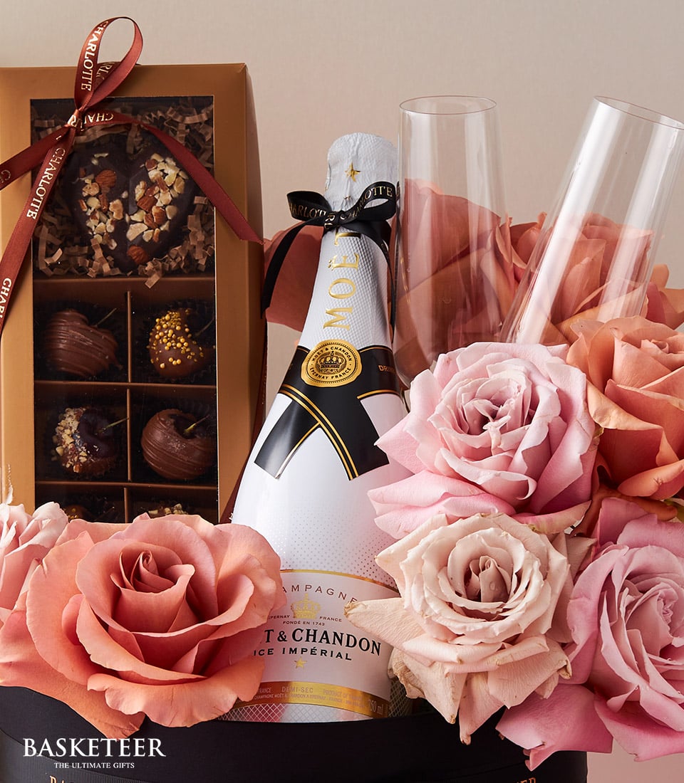 A lavish gift arrangement showcasing a bottle of Moët & Chandon champagne, two exquisite champagne flutes, a decadent box of assorted chocolates, and a stunning bouquet of pink and peach roses. The elegant display is beautifully adorned with a ribbon and box featuring the prestigious 