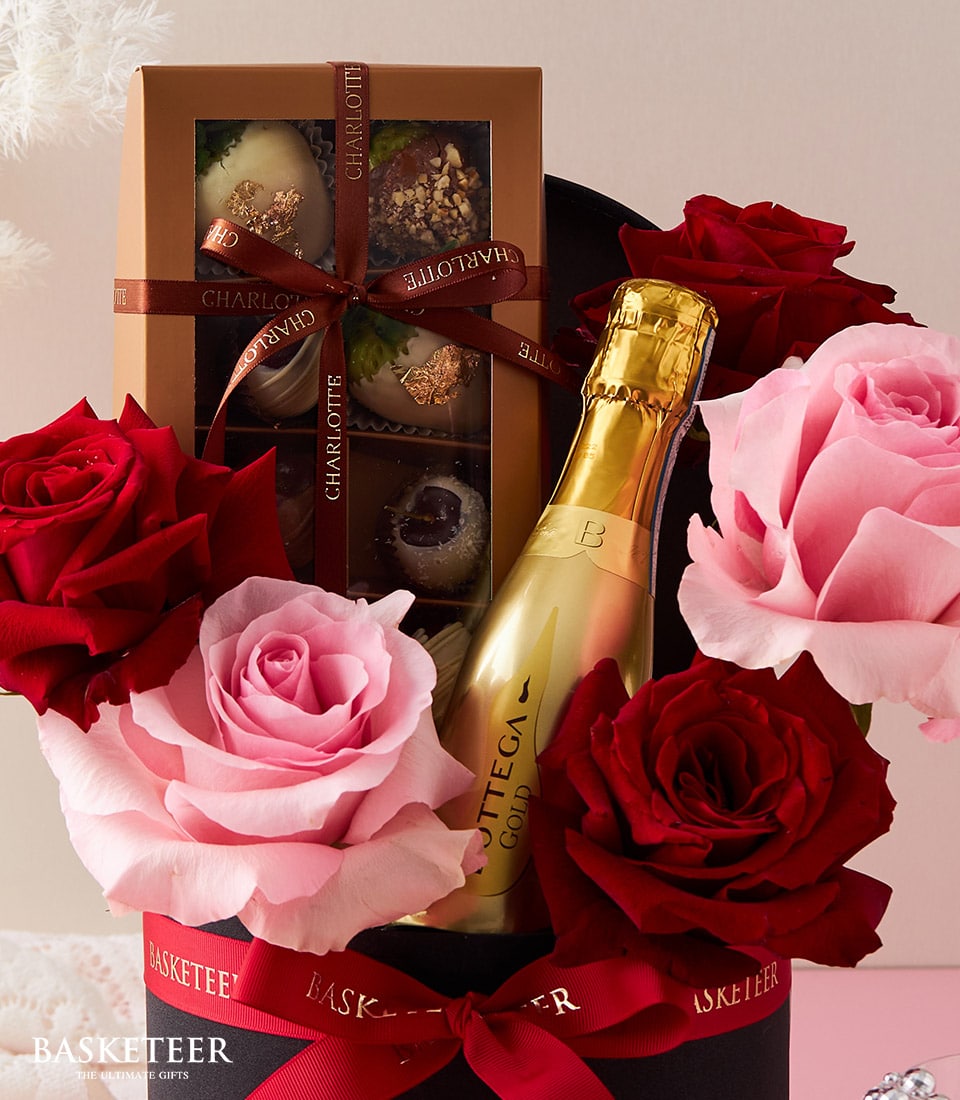 An opulent gift arrangement featuring lush red and pink roses, a gleaming gold bottle of Bottega Gold Prosecco, and a box of exquisite assorted chocolates adorned with an elegant Basketeer-branded ribbon. This luxurious ensemble is carefully presented in a sophisticated black hatbox, proudly displaying the 