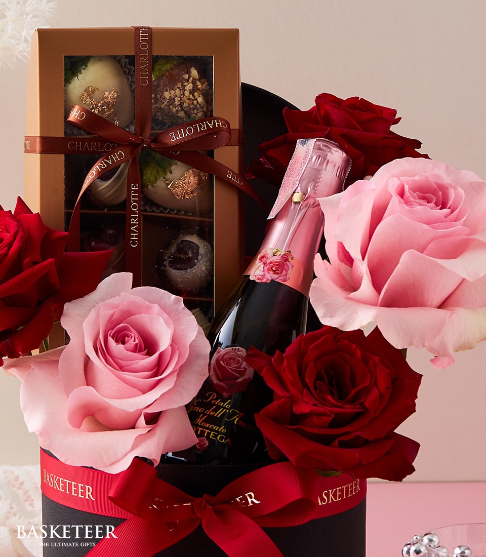 An exquisite gift arrangement featuring a decadent box of assorted chocolates, an elegant bottle of sparkling wine adorned with a chic pink label, and a stunning display of red and pink roses in an opulent black container proudly labeled 