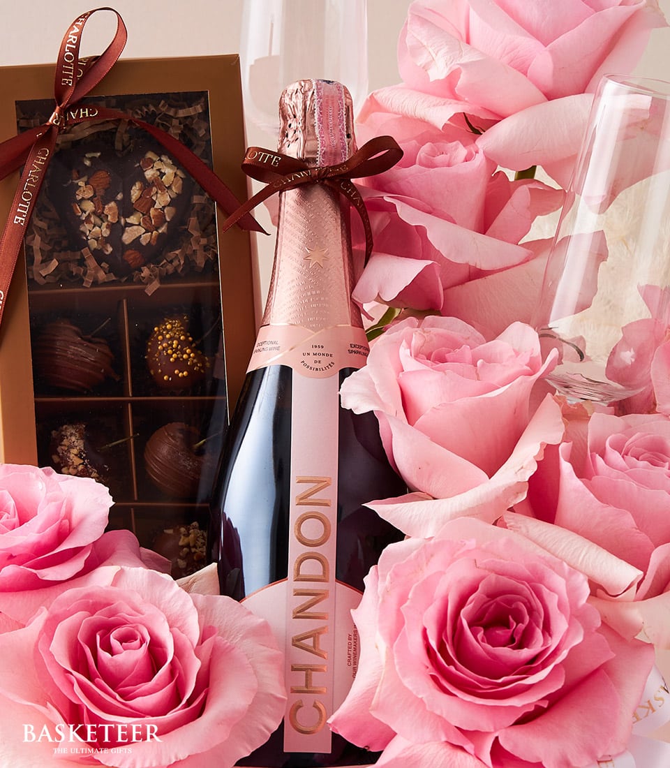 A bottle of Chandon sparkling wine elegantly rests among a bed of luxurious pink roses, evoking an air of celebration and romance. Beside the bottle, a crystal-clear glass awaits the first pour, while a meticulously crafted box of assorted chocolates tempts indulgence. Completing this sophisticated tableau is the Basketeer logo in the bottom left corner, subtly affirming the brand's commitment to opulence and refinement.