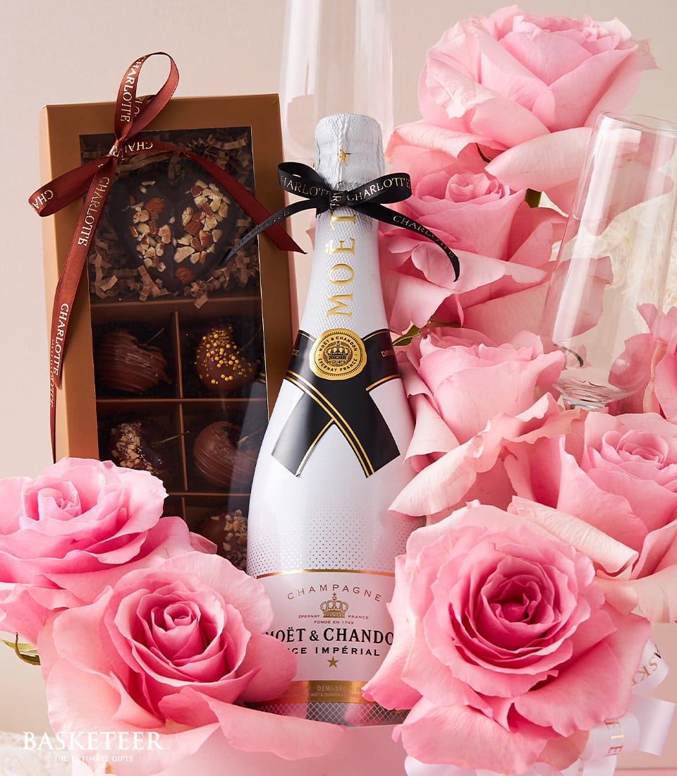 A bottle of exquisite Moët & Chandon champagne, elegantly poised amidst a bouquet of delicate pink roses, exudes an air of opulence. To the left, a charming box of assorted chocolates adorned with the Basketeer logo invites indulgence. Partially visible in the background are two slender champagne flutes, completing this luxurious and inviting arrangement that whispers promises of celebration or romance.