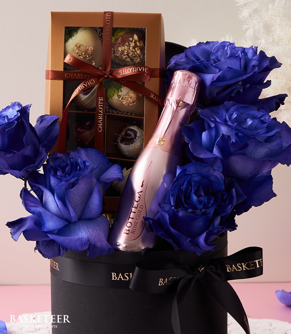 An opulent gift arrangement showcasing a bottle of pink Bottega Rosé Gold sparkling wine, a curated selection of gourmet chocolates, and exquisite romantic blue roses. This luxurious ensemble is artfully presented in a sophisticated black hatbox adorned with a sleek black ribbon imprinted with the 