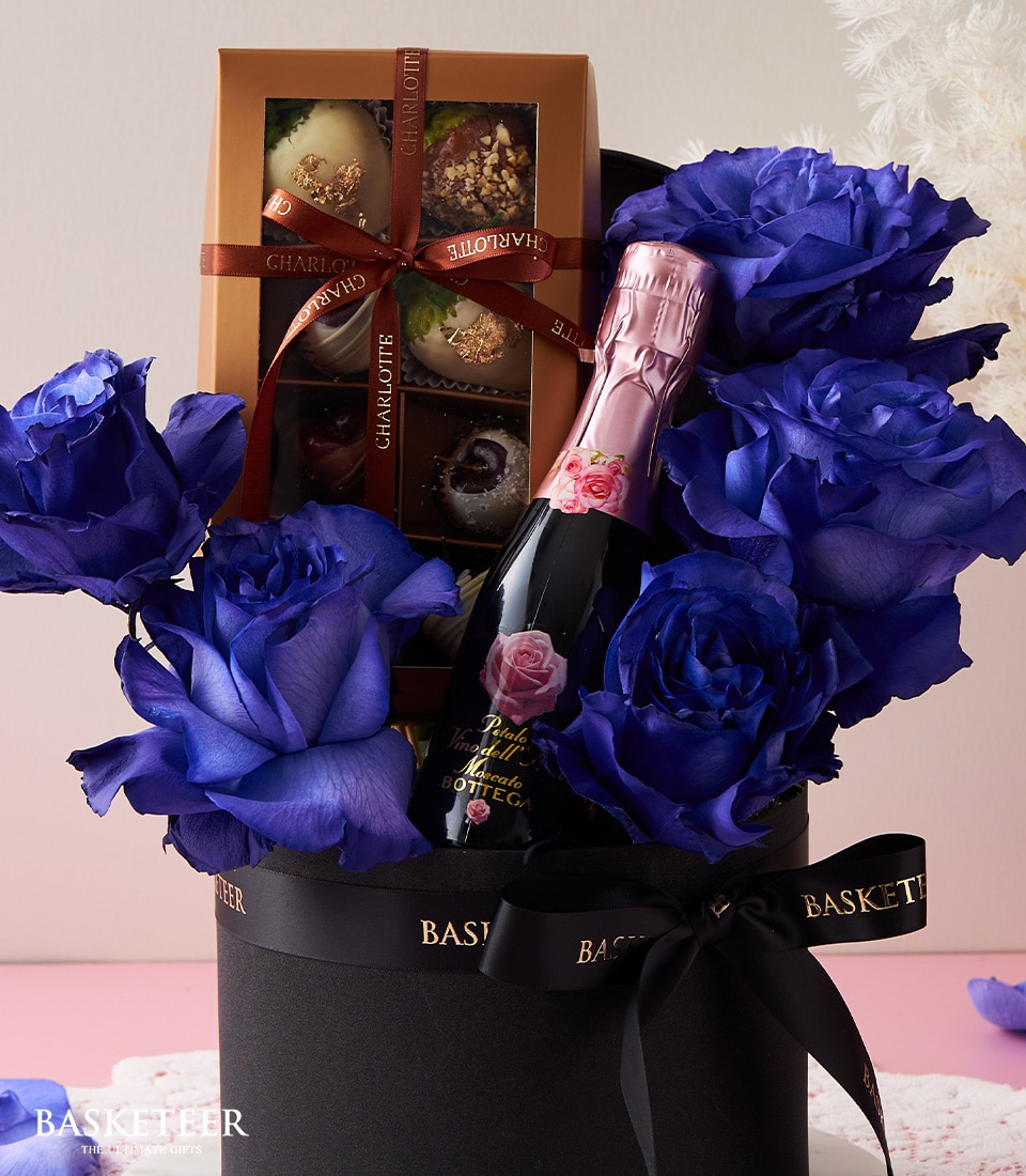 A lavish gift arrangement showcasing a chic black basket adorned with enchanting blue roses, accompanied by a sophisticated pink bottle of sparkling wine and an exquisite box of assorted chocolates. The prestigious 