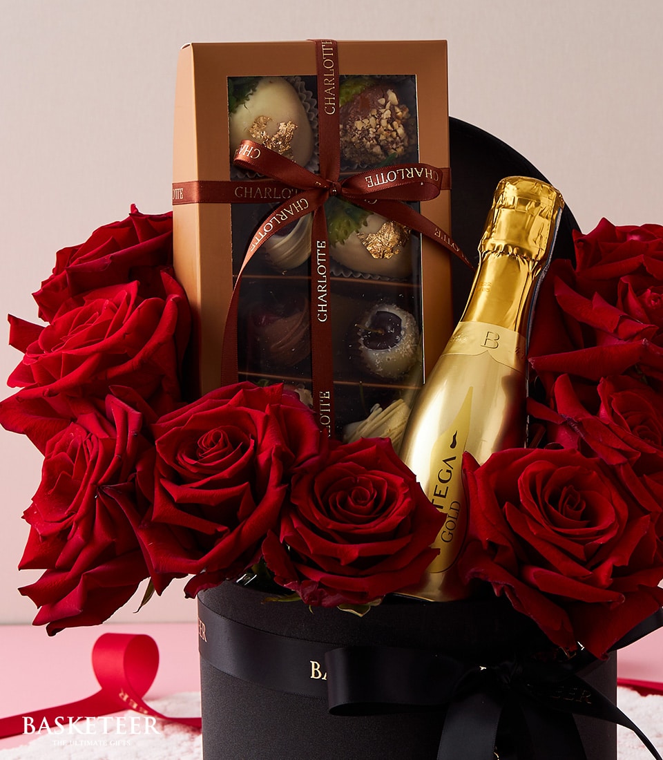 A lavish gift ensemble by Basketeer, showcasing a gold-wrapped bottle of premium sparkling wine, nestled alongside an exquisite box of assorted chocolates in opulent packaging. Radiant red roses add a touch of romance, all artfully arranged in a chic round black box. A sophisticated ribbon adorned with the Basketeer logo completes this luxurious presentation, making it an irresistible indulgence.