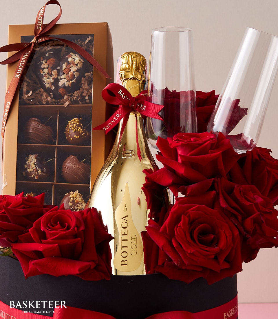 An opulent gift arrangement featuring a shimmering gold bottle of Bottega Gold Prosecco, accompanied by two exquisite champagne flutes. Nestled within the black basket are a luxurious box of assorted chocolates and an array of lush Red Explorer roses. The ensemble is beautifully accented with a crimson ribbon, all presented under the distinguished Basketeer logo, creating an inviting scene of indulgence and elegance.