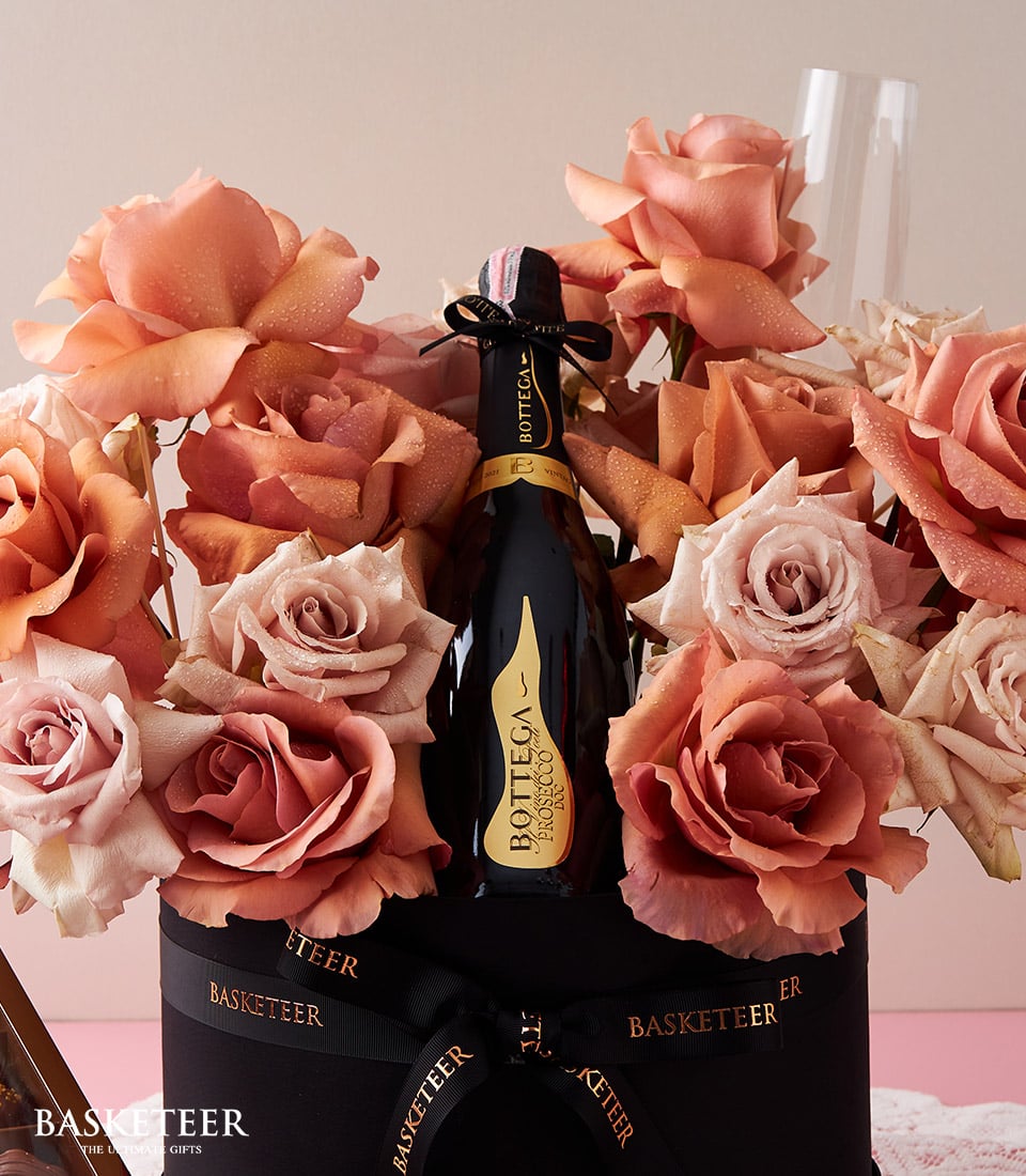 A bottle of Bottega Prosecco graces an opulent black basket adorned with the Basketeer logo, nestled among a lush bouquet of blooming peach and pink roses. The scene is set with two empty champagne flutes nearby, poised for a toast. A box of decadent chocolate-covered treats completes this luxurious ensemble.