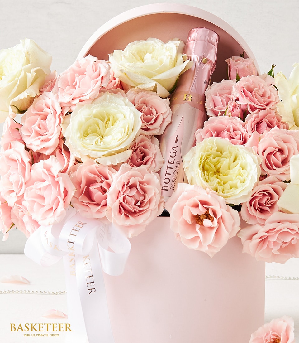A lavish flower arrangement in an elegant pale pink hatbox showcases a stunning ensemble of delicate pale pink, vibrant red, and pristine white roses. Gracefully nestled among the opulent blooms is a sophisticated pink bottle of Bottega sparkling wine. Adding a touch of refinement, a white satin ribbon with luxurious gold text that reads 