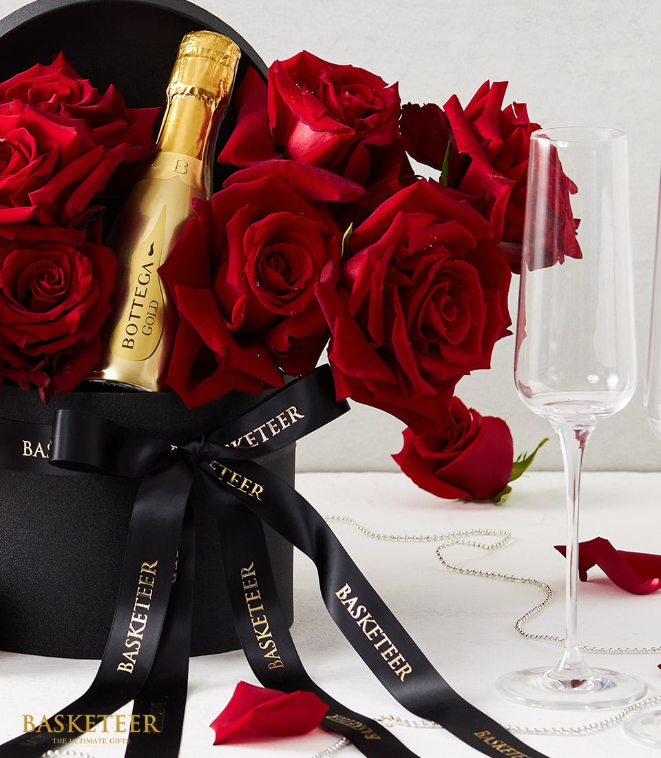 An opulent display showcases a sleek black hat box brimming with velvety red roses, complemented by a lavish gold bottle of Bottega prosecco. The sophisticated box is adorned with a long, elegant black ribbon featuring the 