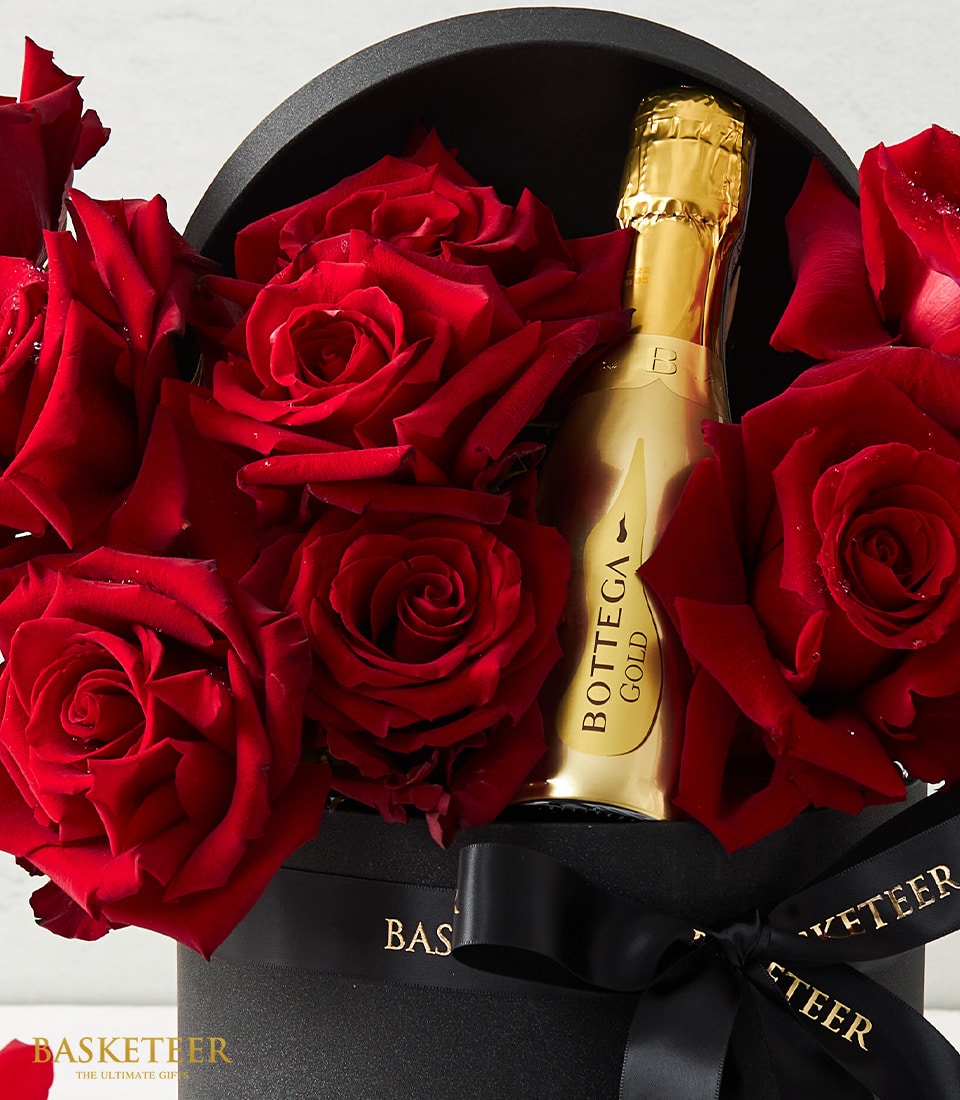 A lavish ensemble showcases resplendent red roses paired with a golden bottle of Bottega sparkling wine, all exquisitely nestled within an elegant black hatbox. The sophisticated box is adorned with a refined black ribbon, subtly embossed in gold with the prestigious 