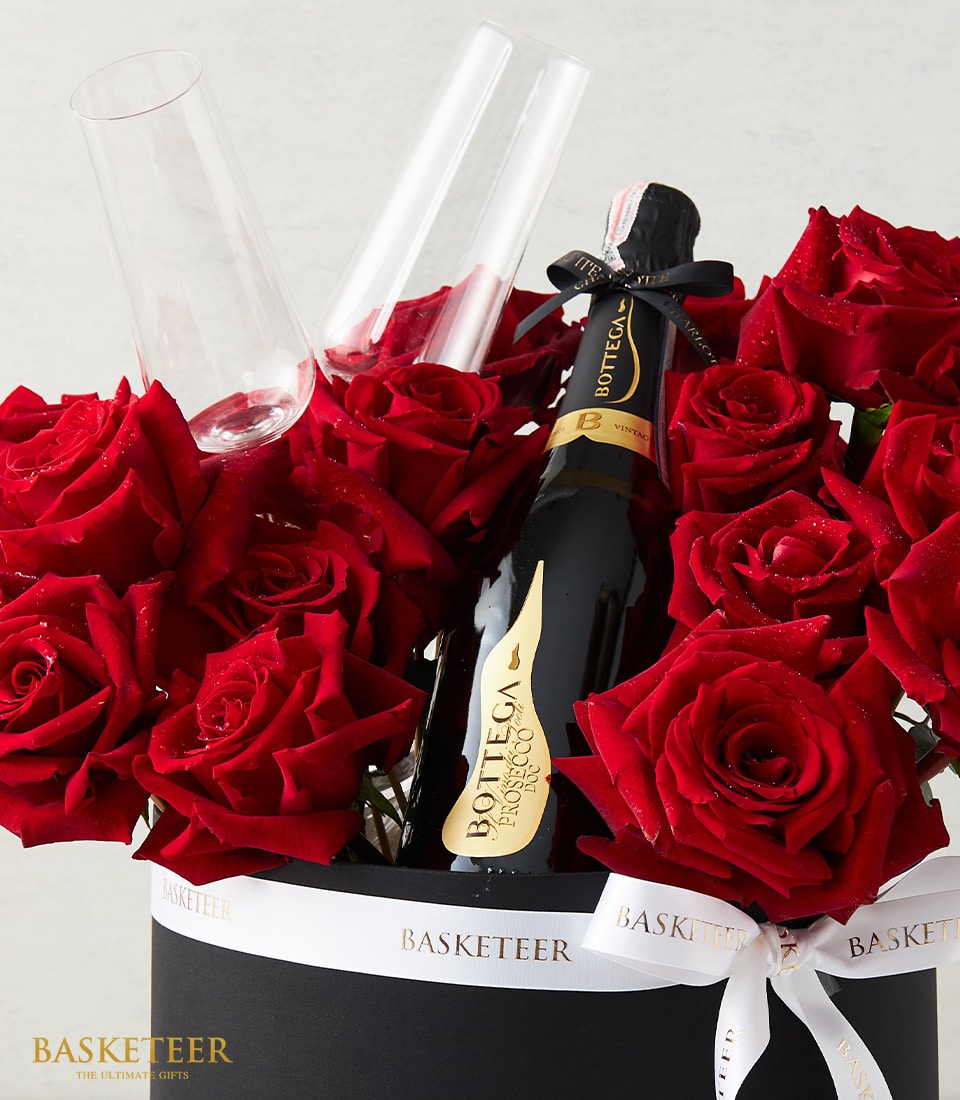 A lavish bouquet of vibrant red roses accompanied by a bottle of Bottega Prosecco, elegantly nestled in a sophisticated black container. Two crystal-clear champagne flutes stand ready for service. Refined white ribbon emblazoned with the 