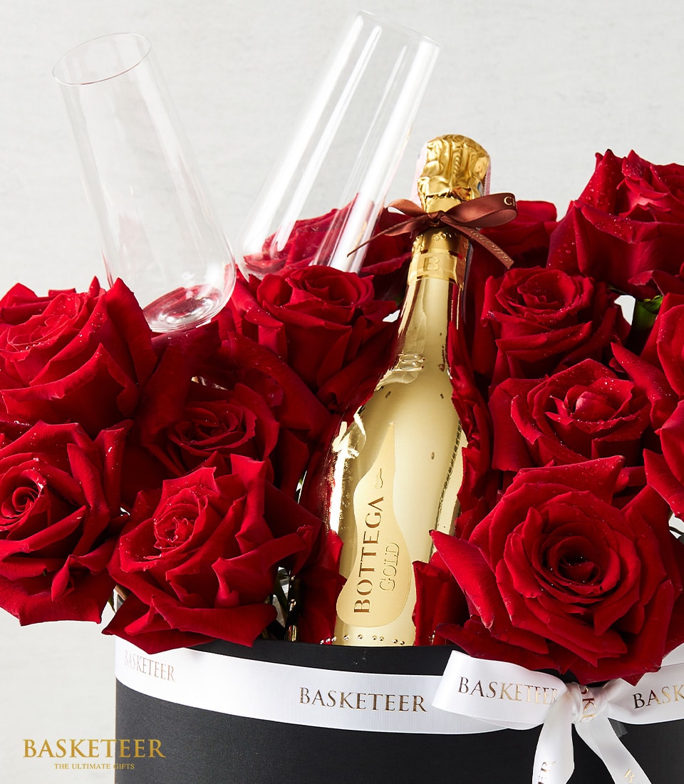A sumptuous bouquet of velvety red roses elegantly cradles a golden bottle of Bottega Gold Prosecco. Nestled among the lush petals, two sophisticated champagne flutes await to enhance your celebration. This opulent arrangement is tastefully presented in a sleek black basket adorned with the distinguished 