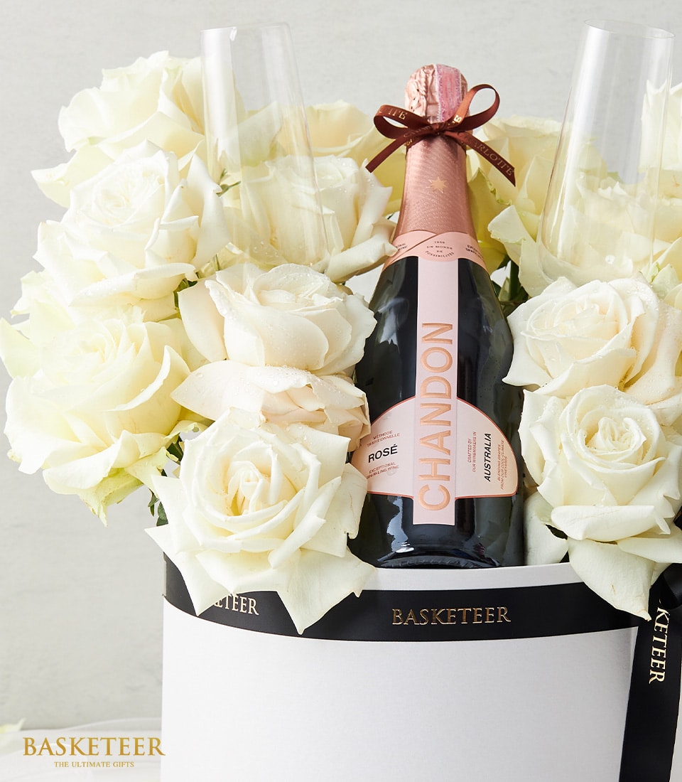 A bottle of exquisite Chandon rosé champagne, elegantly nestled amid luxurious white roses, is flanked by two crystal champagne glasses. The entire arrangement is presented in an opulent white box with sophisticated black trim, proudly displaying the 