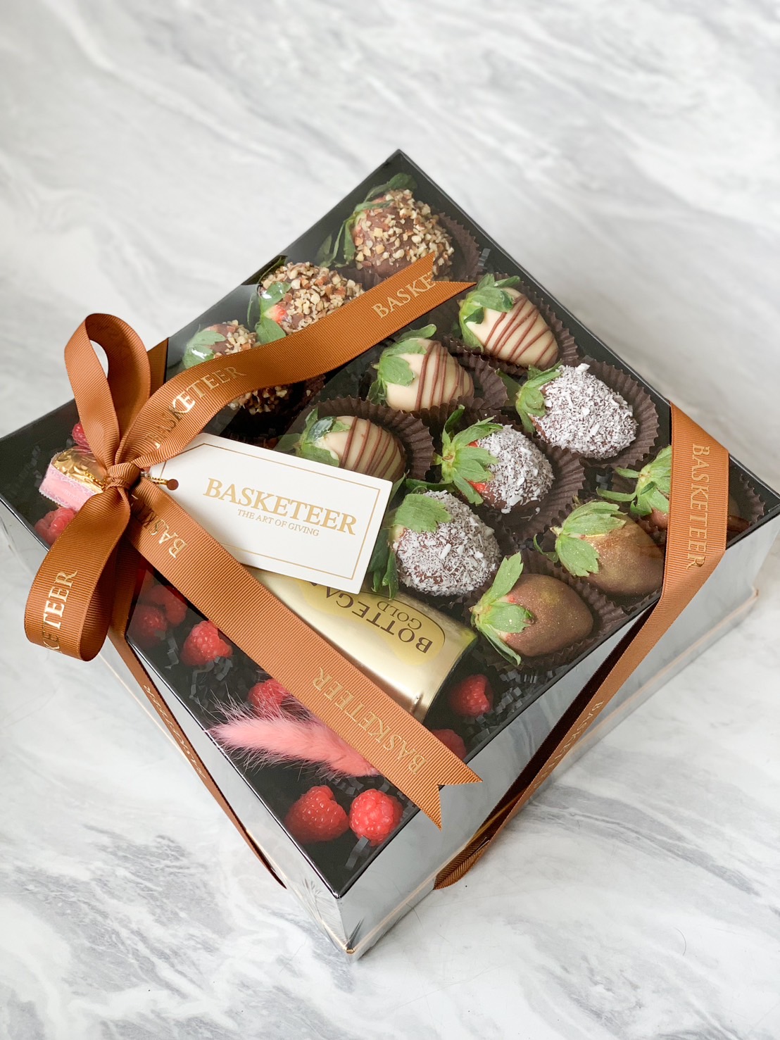 An opulent black Basketeer gift box showcases a sumptuous array of chocolate-covered strawberries, a petite bottle of Bottega Gold prosecco, and fresh raspberries. Each element is adorned with intricate gold pearls and complemented by a luxurious red rose and elegant ribbon bearing the Basketeer logo. The backdrop is draped in lustrous red and silver satin fabric, embellished with shimmering beads and an exquisite card, creating an inviting scene of indulgence.