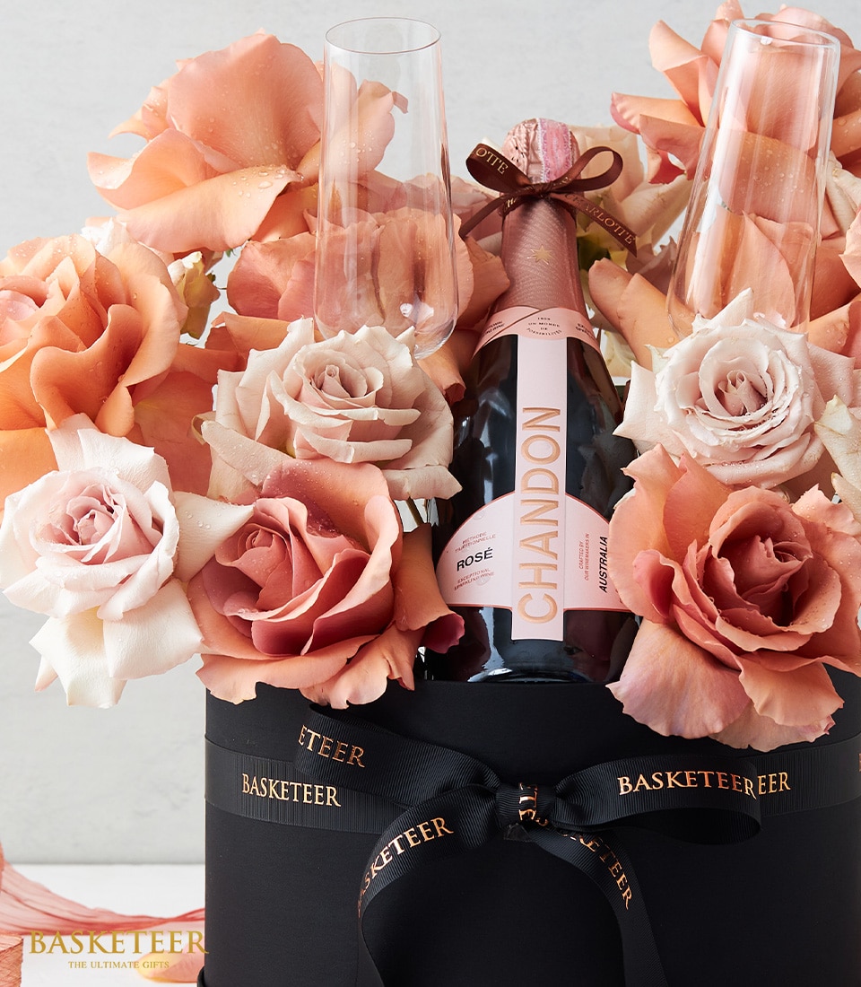 A lavish gift arrangement designed to impress, featuring a premium bottle of Chandon Rosé elegantly wrapped in a chic ribbon, nestled amidst an opulent array of peach and white roses. Two exquisite champagne flutes are gracefully placed within the stunning floral display. The entire presentation is impeccably housed in a sophisticated black box adorned with the distinguished 