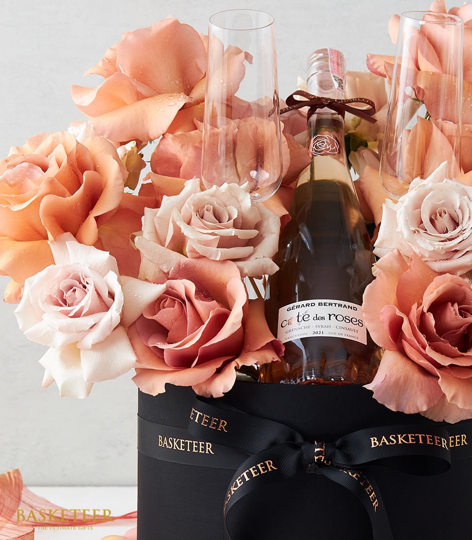 A luxurious gift arrangement showcases a bottle of Gérard Bertrand Côte des Roses rosé wine, elegantly cradled among peach and cream roses. Behind the bottle sit two pristine champagne flutes, ready to elevate any celebration. The entire scene is presented in a sophisticated black box, adorned with an exquisite 
