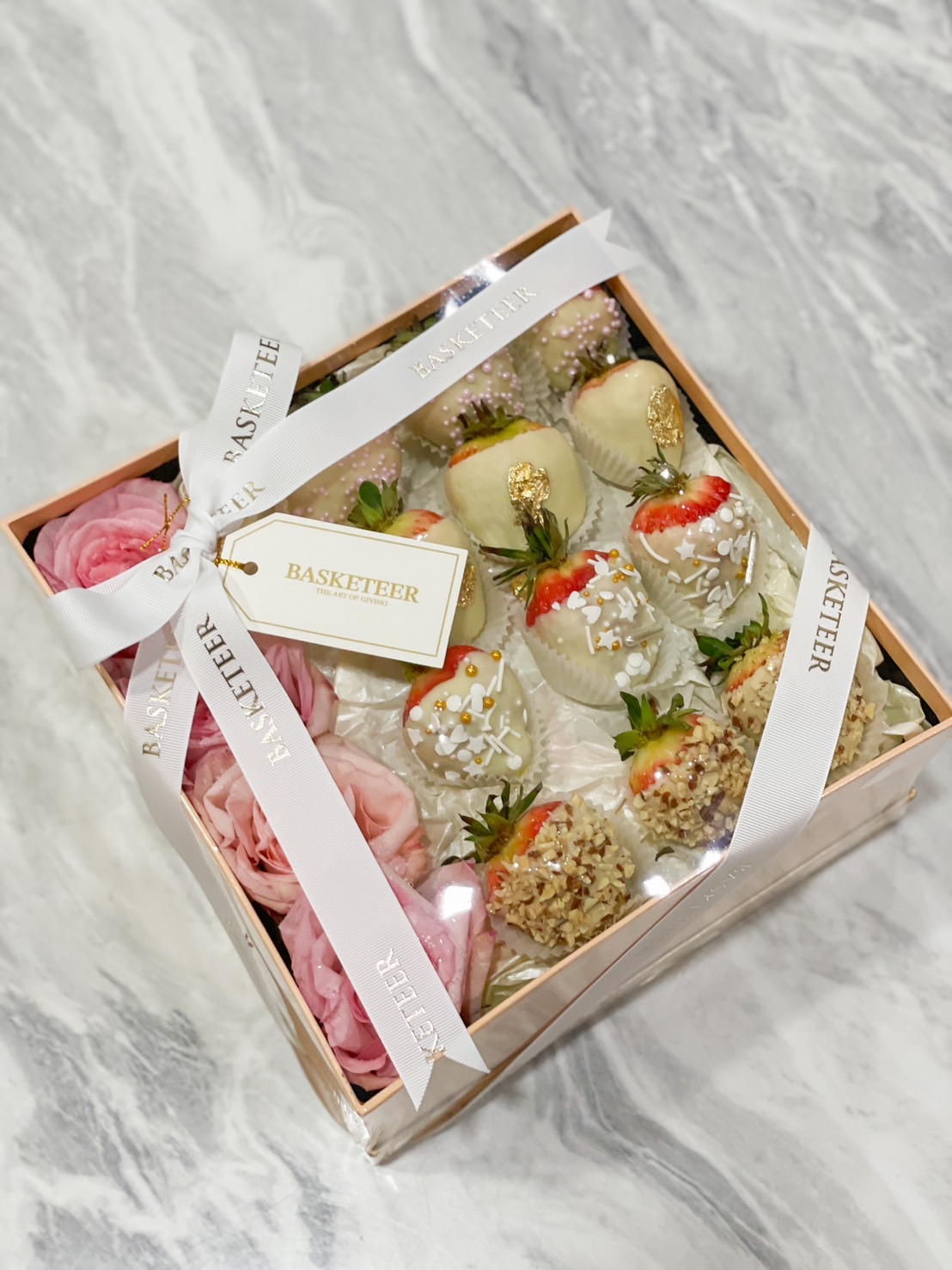 Elegant gift box featuring white chocolate-covered strawberries with gourmet toppings, beautifully arranged with fresh pink roses and wrapped with a white satin ribbon.