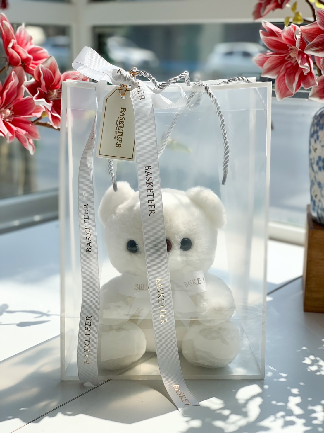 A beautifully packaged white plush teddy bear inside a clear luxury gift bag with a Basketeer ribbon, perfect for gifting.