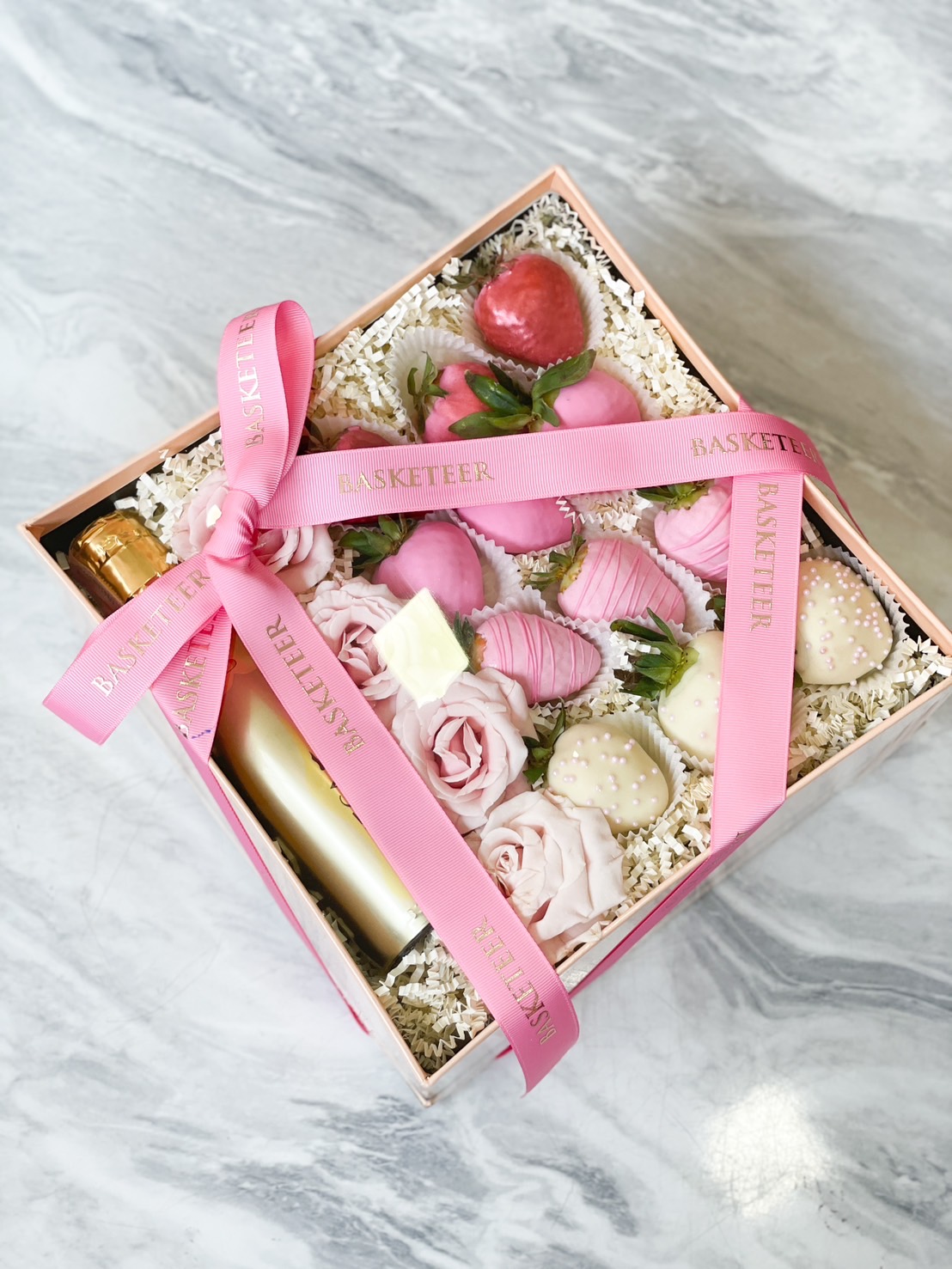 A luxurious pink-themed gift box featuring chocolate-covered strawberries, fresh pink roses, and a bottle of sparkling wine, elegantly wrapped with a pink Basketeer ribbon.
