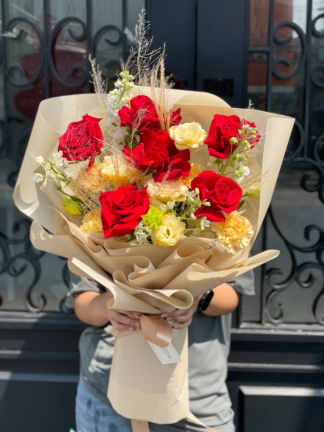 A gorgeous bouquet of red roses and soft yellow blooms, beautifully arranged with dried floral accents and wrapped in luxurious beige paper, creating an elegant rustic floral arrangement.