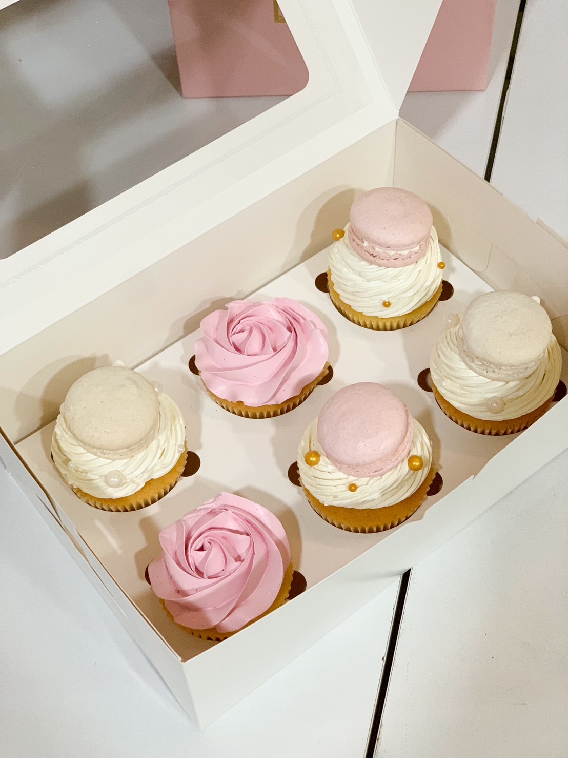 A box of six elegant cupcakes featuring rose-shaped buttercream swirls and macarons, decorated with delicate pearls and gold accents.