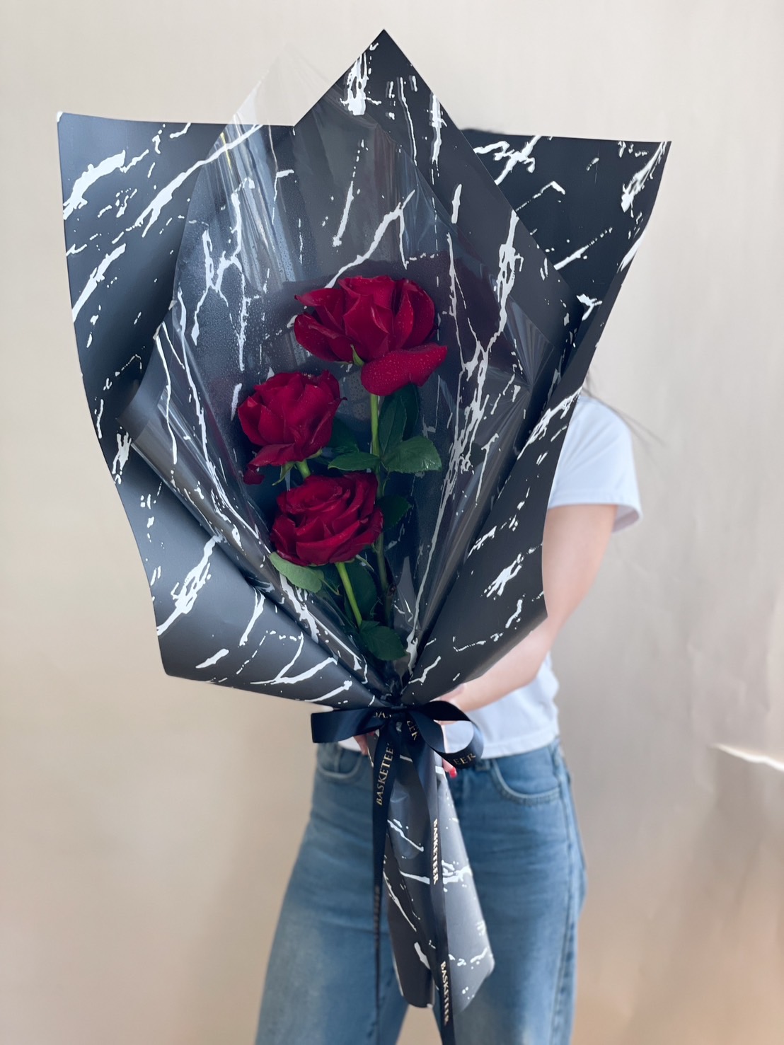 A stylish bouquet of three deep red roses, wrapped in black marble-pattern paper with a satin ribbon, held by a person in a white shirt.