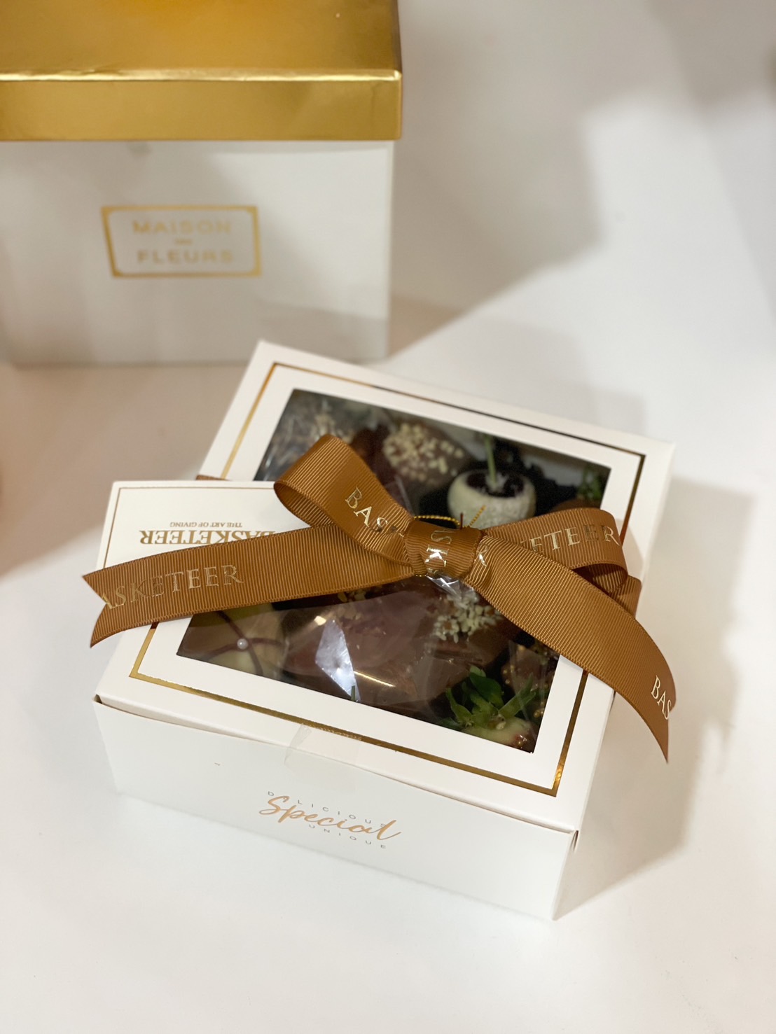 Elegant white and gold gift box of gourmet chocolate-dipped fruits, tied with a luxurious brown satin ribbon.