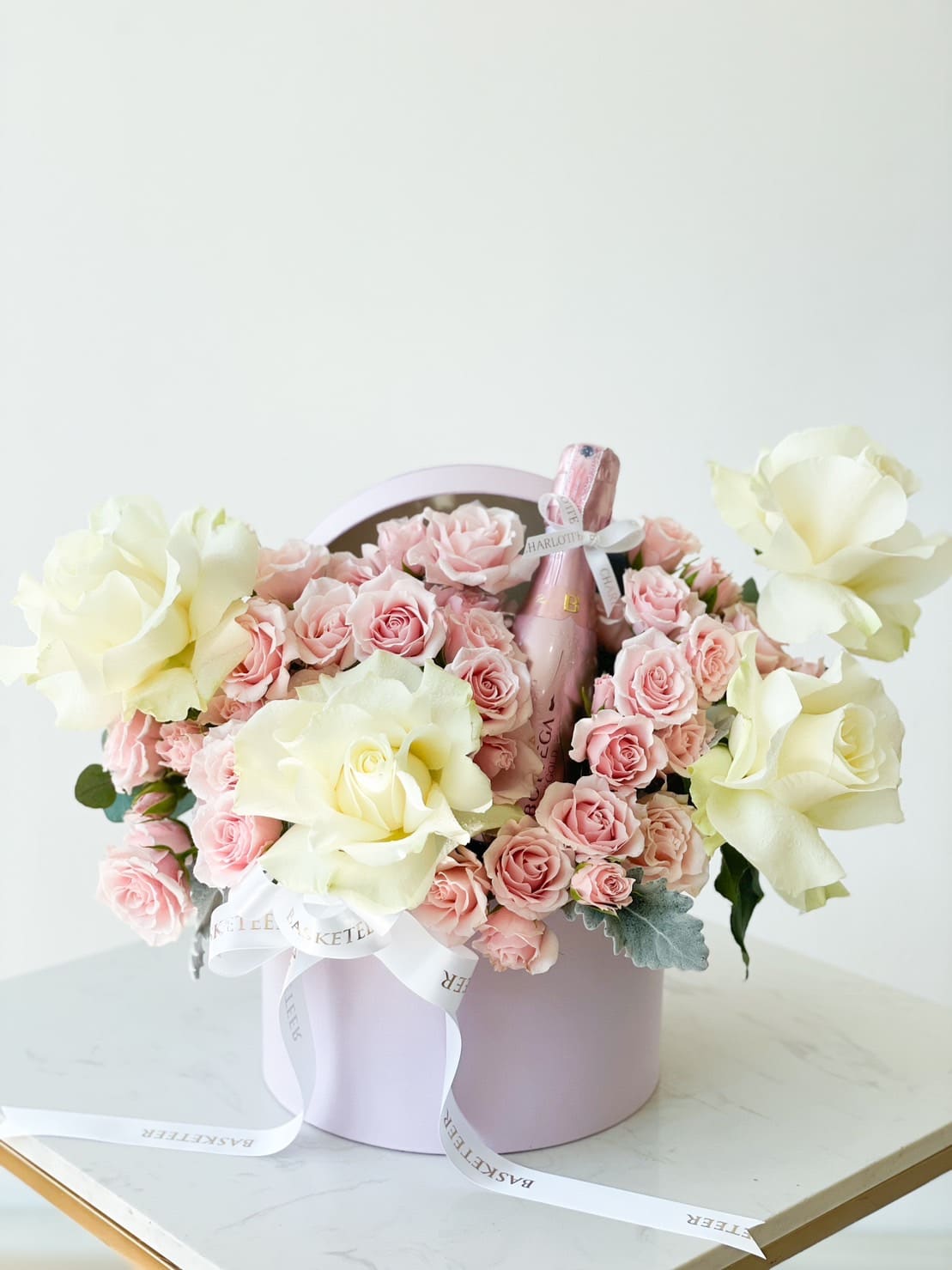 A pastel pink hatbox filled with an ensemble of soft pink, cream, and red roses surrounds a pink bottle of sparkling wine. White ribbons with text "MASTELI" adorn the flowers. The arrangement is set against a plain white background.