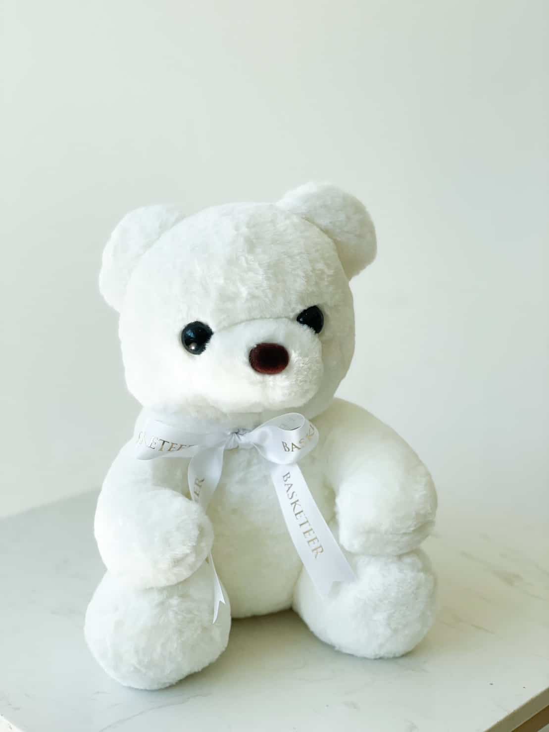 A white teddy bear sits on a light-colored surface against a plain background. The bear has black eyes, a dark nose, and is adorned with a white ribbon around its neck with the word "BASKETIER" printed on it, completing the charming ensemble.