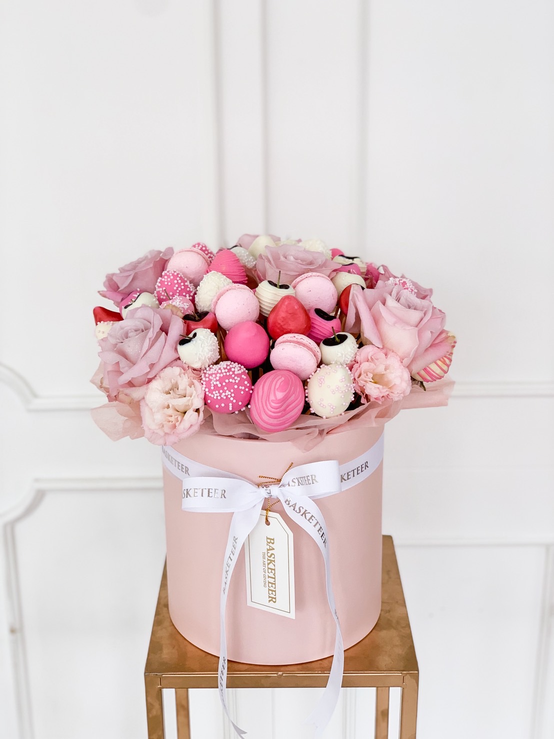 Pink round gift box filled with roses, pink chocolate strawberries, macarons, and candy pops